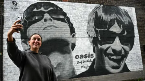 Getty Images Fan Emily McShane takes a photo of a new street artwork depicting Liam and Noel Gallagher of Oasis, created by Manchester-based street artist Pic.One.Art. on the side of the Sifters Record store in Burnage, a suburb of Manchester, northern England on August 27, 2024