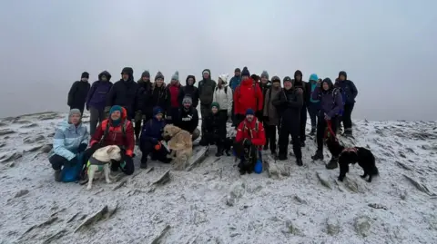 The Newcastle Hiking Group The Newcastle Hiking Group - a group of hikers stand together a top of a fell summit. The group are wearing winter warm clothing and outdoor gear. There are a few dogs. The cloud is thick grey and there is small layer of snow on the ground. 