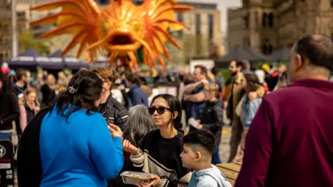 Bradford Council Families enjoying themselves in a festival setting. In the foreground is a family chatting and enjoying some street food. In the background is a huge puppet fish making its way across a city square