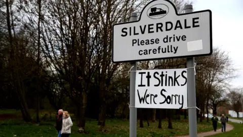 Reuters A road sign reading: "SILVERDALE - Please drive carefully." Underneath, a hand-written sign reading: "It stinks - we're sorry" has been tied to the signpost.