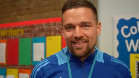 Jamie Niblock/BBC Ben Andrews smiles at the camera while standing in front of several school notice boards. He has short dark hair with a dark beard. He is wearing a blue jacket.