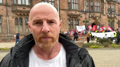 Matthew Reid stands in front of protestors outside Coventry Council House - a large sandstone building. They wave flags and hold up placards out of focus in the background. Matthew has a shaven head and a ginger beard. He wears a white t shirt and a black coat.