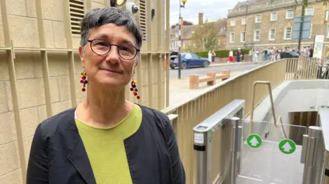 Qays Najm/BBC Head and shoulders image of Katie Thornburrow. Katie has short grey hair, is wearing earrings and rounded glasses, a green top and black overshirt. She is stood near the entry to toilets.