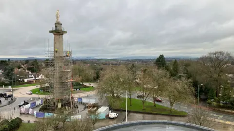 A tall column covered in scaffolding with a statue, Lord Hill, on top made of it. A roundabout can be seen behind it with hills visible in the background.