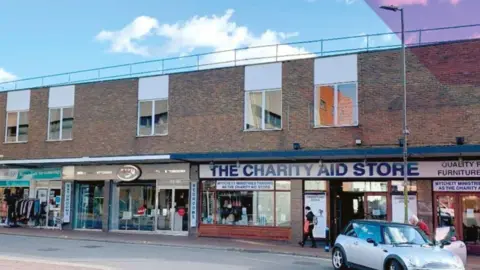 A row of shops in Camberley, including a charity shop. A mini is parked at the kerb and a pedestrian is walking past.