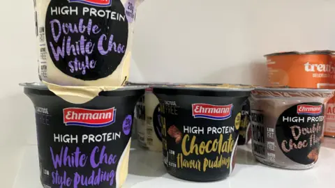 Pots lined up on a shelf of Ehrmann High Protein White Choc Style Pudding, Ehrmann High Protein Chocolate Flavour pudding, Ehrmann High Protein Double Choc flavour and Ehrmann High Protein Double White Choc Style.