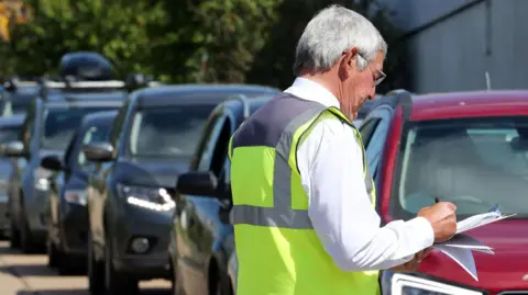 A man with grey hair wears a hi-vis vest as he checks through some paperwork at a recycling centre. There is a queue of cars next to him.