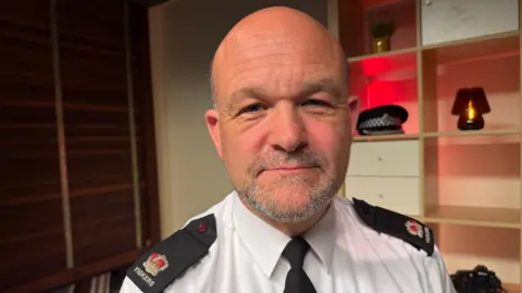 A head and shoulders photo of Phil Vickers, a man with a bald head, close-cropped grey hair at the sides and a close-cropped beard. He is wearing a smart white shirt with black shoulder epaulets, carrying his rank in ther form of a red and gold crown, and his surname, Vickers, and a black tie. He is in what looks like a study with shelves behind him, which hold a police cap, camera and lamp.