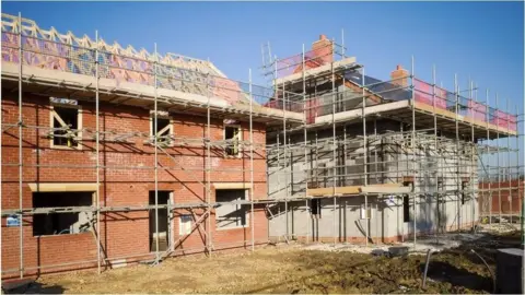 Getty Images Partially constructed residential buildings surrounded by metal scaffolding at a housing development site. The red-brick homes are in various stages of construction, with exposed roof structures and unfinished walls visible. The foreground shows bare earth and construction debris, while the background features a clear blue sky. The image illustrates new homes being built on a development site.