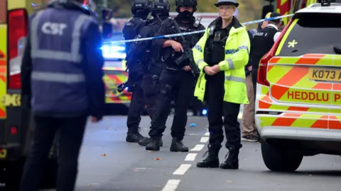 Police officers, some of whom are armed response officers, stand near police cars and other emergency vehicles behind a cordon