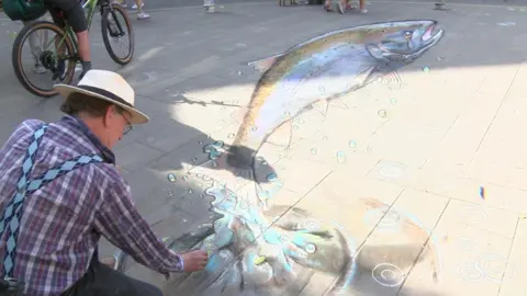 A large chalk salmon being drawn by the artist Julian Beever, who can be seen on his knees.