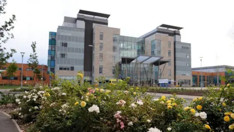 The front of Peterborough City Hospital. It has light brown cladding and multiple windows. Pink, yellow and white flowers are in the foreground.