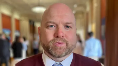 A close-up of a man's face. He is bald with a light-coloured, grey-flecked beard. He is wearing a white shirt, blue tie and a burgundy jumper or sweater. He is standing in the room. In the background, out of focus, there are other people milling about and talking. 