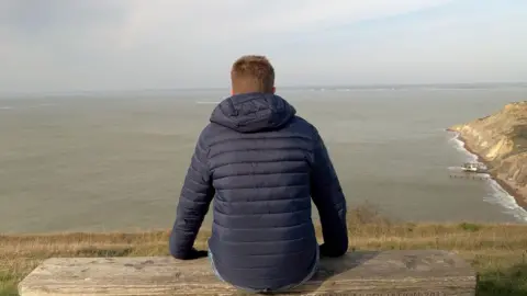 A man is sat on a bench on a hilltop. He is wearing a blue coat and blue jeans. There is grass all around and the sky is overcast. The sea is below. 