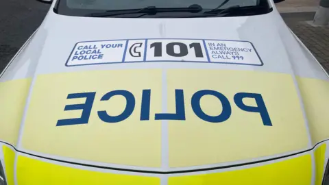 The bonnet of a police car. It is yellow and white. It has "police" written in blue block capitals backwards. It also has a large sticker on the bonnet urging people to call 101 or 999. 