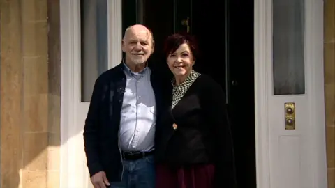 A man and woman stood together outside their black front door. The man is bald and has a grey goatie beard. He is wearing jeans, a blue jacket and blue shirt. The woman has short, brown hair and is wearing a dark top with a blue and yellow patterned shirt