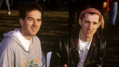 Getty Images Jim "Bob" Morrison with short dark hair wearing a red headband, a black leather jacket and a white T-shirt, sitting alongside Les "Fruitbat" Carter, with a grey top, white T-shirt and medium-length dark hair.