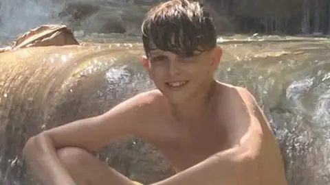 Family photograph Jools sat in a river or stream of water smiling at the camera. He has brown hair which is swept across his forehead