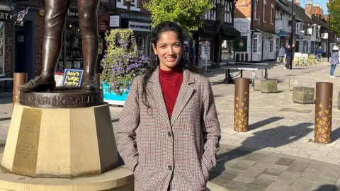 Lakshmi Balakrishnan stood wearing a brown coat over a red sweater. Behind her lies the main street of Stratford-upon-Avon with the base of a statue of William Shakespeare.