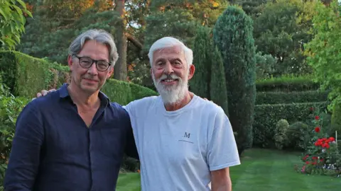 Contributed Two men standing in a garden with their arms around each other. One the left is a grey haired man wearing black-rimmed glasses and a blue shirt. On the right is a man with grey hair and a grey beard wearing a pale T-shirt marked on the left side of the chest with the letter "M".