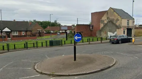 A roundabout connecting High Street and Elemore Lane in Sunderland. A black car is travelling away from the camera. A number of brick buildings can be seen to the side of the road.