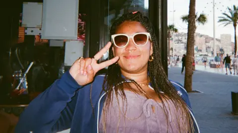 Beth Doherty Beth Doherty, photographed on a grainy film camera. She is wearing sunglasses and is holding her fingers up in a V symbol, while smiling. 