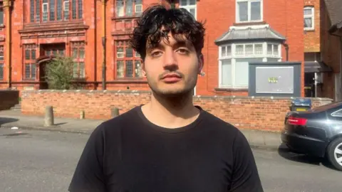 A mid shot of a man with short curly, black hair. He is standing on a residential street with a red brick building behind him. He is looking directly into the camera and is wearing a black t-shirt. 