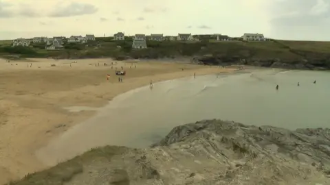 A picture overlooking a sandy beach which has people on it. The sea is blue and at the forefront of the image is a rocky cliff. In the distance is green grass and houses dotted along it. In the distance, at the centre of the beach, there is a red and yellow flag.
