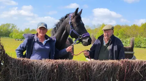 Angela Winter Angela Winter and "Grandad Bob" standing either side of a black horse. Angela is wearing a purple polo shirt, a purple coat unzipped and a green cap. Bob is wearing a green polo shirt, a blue coat unzipped and a green flat cap. The horse is wearing a blue and purple head collar. They are standing behind a cross country fence in a field. 
