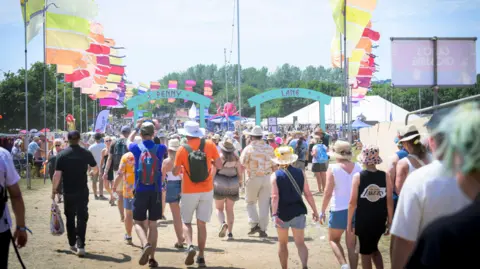 Getty Images A crowd of people walking through a festival site are pictured from behind. Many of them are wearing hats and summer clothes. Rows of brightly coloured flags can be seen flying above them. 