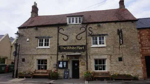 Outside the White Hart pub in Wolvercote on a cloudy day. There are flower beds in front of the building and a wooden sign by the door. Two wooden benches are places on each side of the door.