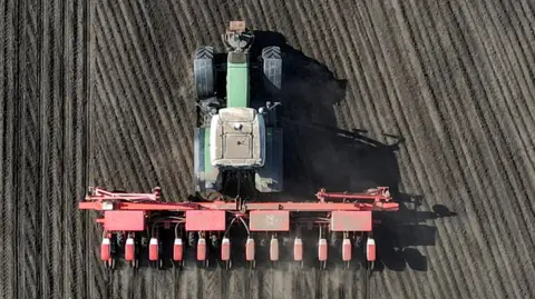 An aerial view shows a green tractor pulling a wide red planting or seeding machine across a large field, creating neat, evenly spaced rows in the soil. The machinery is moving forward in straight lines, leaving darker, freshly worked earth behind it in a carefully patterned grid.