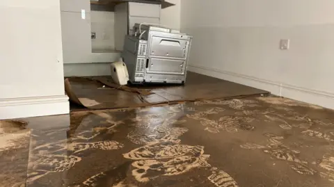 A room which shows a muddy, wet floor with footprints. There is a line about waist-high on the walls which shows how high the water went. In the background there is a kitchen applicance.