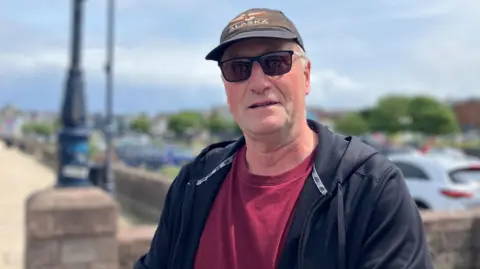 A man is standing in a path with cars parked behind him. He's wearing a cap and sunglasses with a jacket and a maroon top.