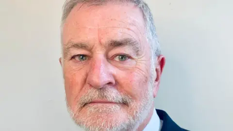 A man looks at the camera, he has a white, short beard and short white hair. He appears to be wearing a suit and shirt.