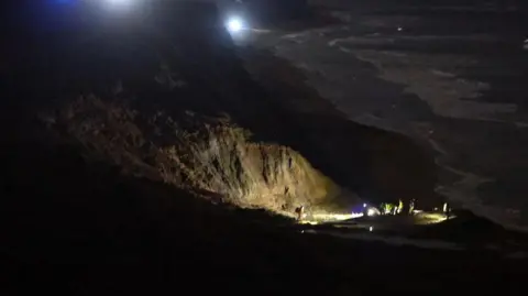 Island Echo Outline of cliffs on Isle of Wight at night with beach and sea to the right - lights can be seen on the beach where a rescue of two people stuck in mud is underway.