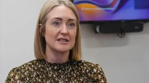 PA Media Esther Ghey, with shoulder length blonde hair and wearing a flowery top with a silver necklace, speaks to the camera