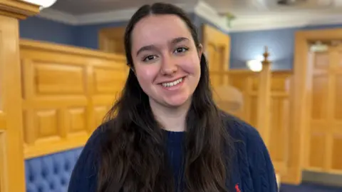 Alex Cowley has long black hair which is half up and half down. She is wearing a navy jumper and standing in a room panelled in pine wood with blue at the top part of the wall. To the left the top of banquette seating in blue leather can be seen.
