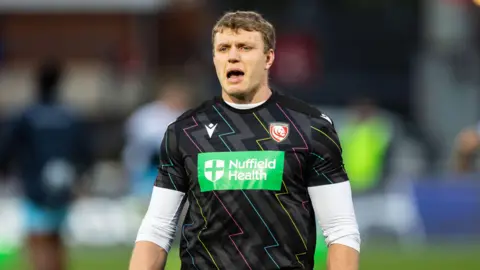 Ollie Thorley standing on the pitch during a pre-game warm-up with his arms by his side and his mouth open
