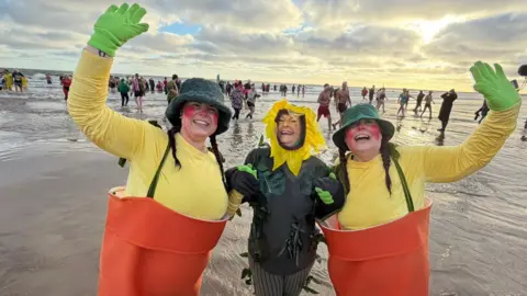 Three women standing in the sea. The woman in the middle is dressed as a sunflower and smiling. The two on either side of her are dressed as plant pot with braces holding up the pots. They have bright face paint on and are holding their arms in the air. People can be seen behind them in the sea. The sun is rising and the sky is covered in clouds.