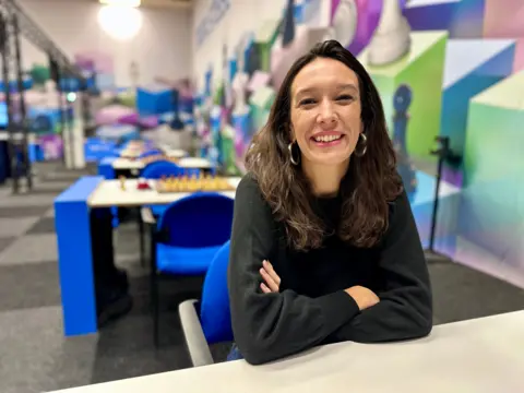 A woman in a dark sweater is seen sitting at a table. She is smiling, with dark long hair and silver hoop earrings. Behind her is a row of tables with chess boards.