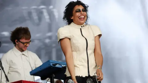 EPA A woman is standing on stage in a white top laughing, with her tongue sticking out between her teeth. She has dark, black eyeshadow all around her eyes. Behind her is a man playing synths.