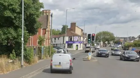 A junction with traffic lights on Dereham Road. Vehicles are travelling in both directions along the road, which is bordered by trees, a modern church building and businesses.