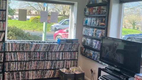 Rachael McMenemy/BBC Shelves packed with DVDs on another wall of the shop. There is also a TV on another wall with a film playing.