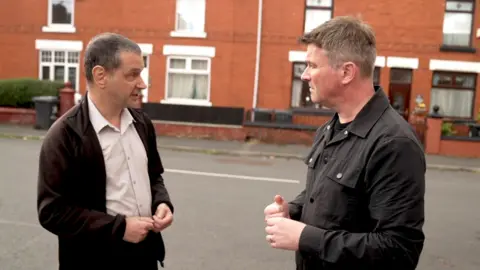 Adrian speaks to Ed Thomas both men wear black coats they are standing on a street with terrace houses.