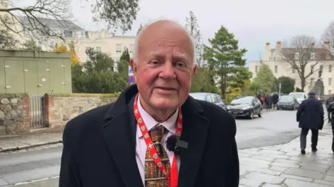 BBC Chris Le Tissier in a suit and colourful tie, smiling on a street with cars and pedestrians behind him