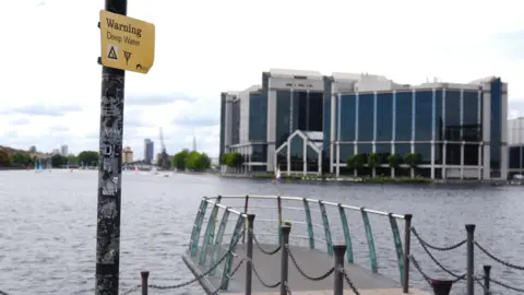 PA Media Warning sign for deep water beside a riverside walkway and viewing platform, with office buildings across the water.