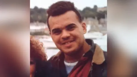 Yeti Television/BBC Cymru Wales Archive photograph of Tyrone France, who smiles at the camera. He has brown curly hair, brown eyes and stands on a bridge outside. He wears a blue t-shirt, red shirt and a coat. 