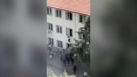 Reuters A group of people stand on the pavement trying to catch people jumping from a first-floor window in a three-storey building.
