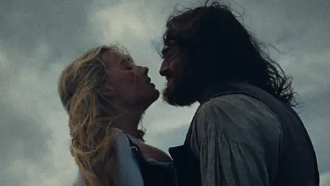 Warner Bros. Ent. Character Cathy and Heathcliff, both with windswept hair and in period costume, share a passionate embrace set against a moody sky